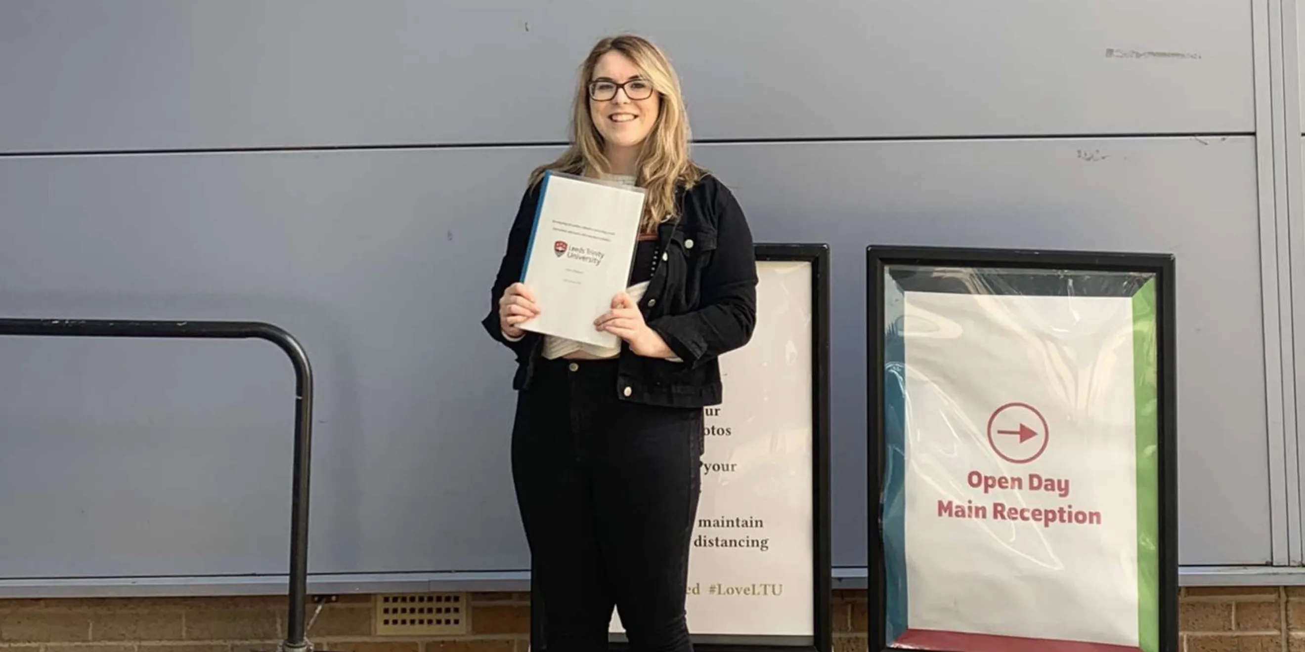 Blonde girl with black glasses in black denim jacket and jeans holds dissertation on white paper.