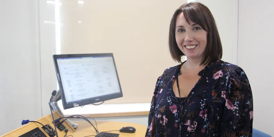 Female with short brown hair stands next to computer.