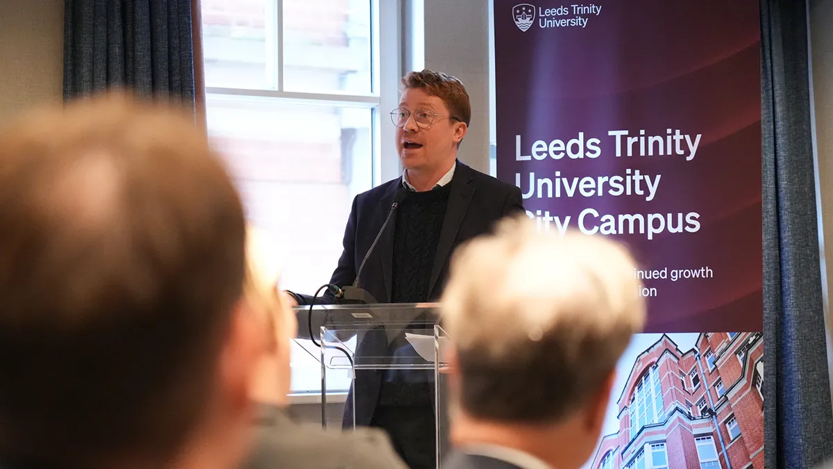 A male councillor speaking in front of an audience with a banner that reads Leeds Trinity University City Campus behind him..