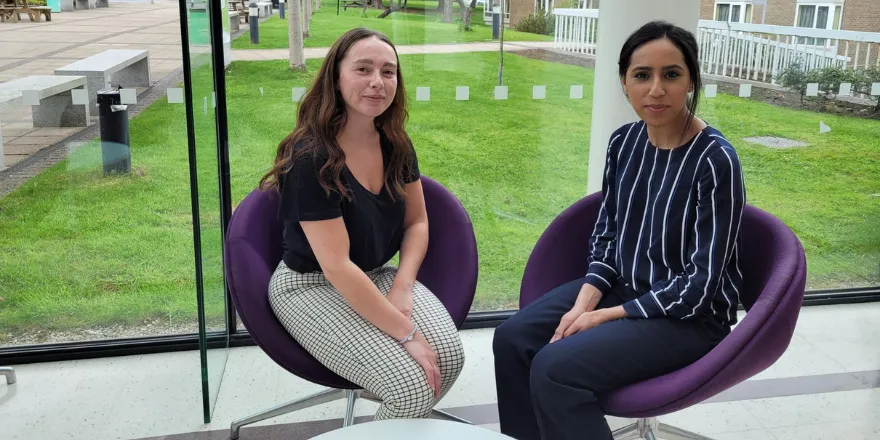 Two female students sit on purple chairs in front of atrium window.