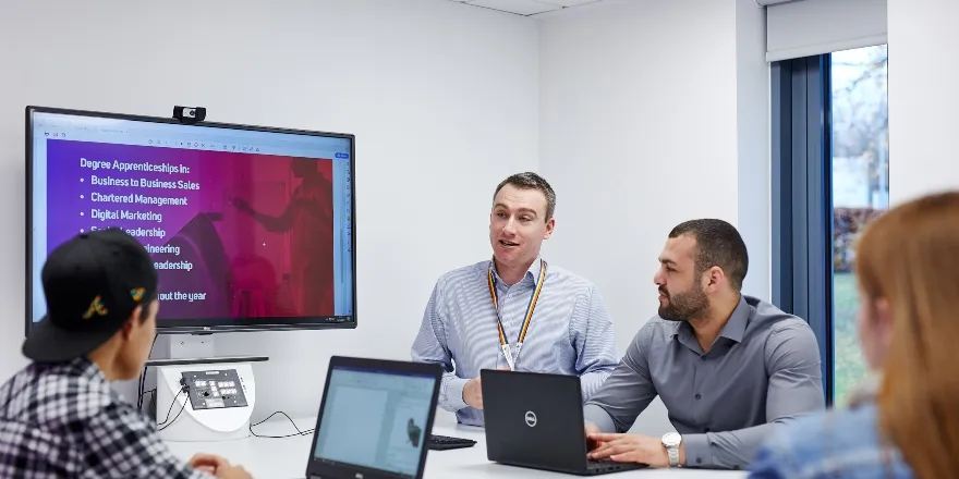 Men in shirts with laptops at white desk and large TV screen.