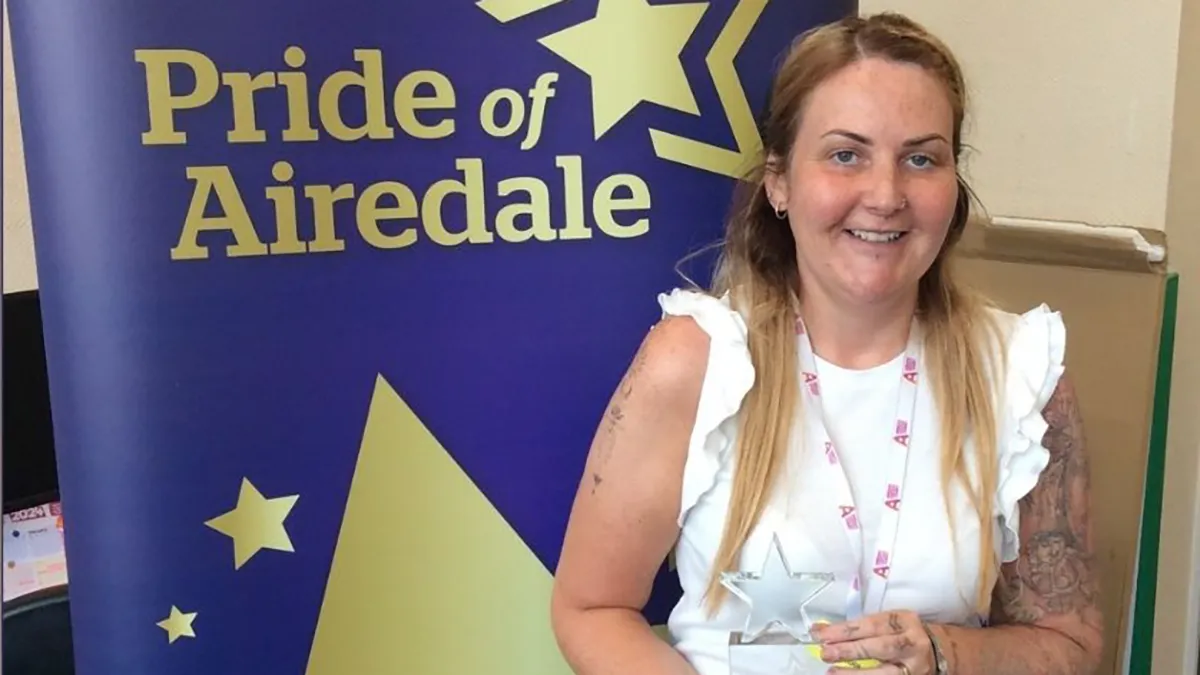 A mature student posing with an award against a purple and gold background that reads Pride of Airedale..