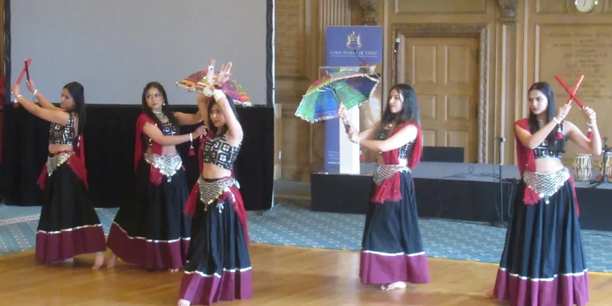 Group of five female dancers stand in front of stage dancing with fans.
