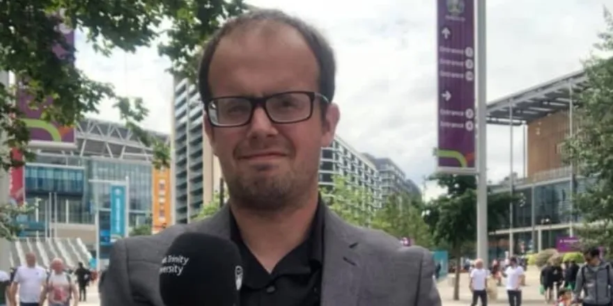 Male student with glasses stands in front of Wembley Stadium wearing grey suit jacket and black shirt holding a black microphone.