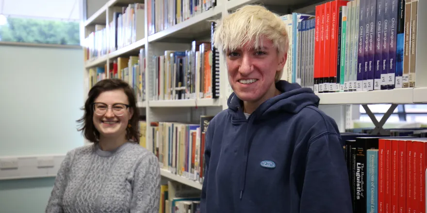 Two female students stand in front of bookshelf.
