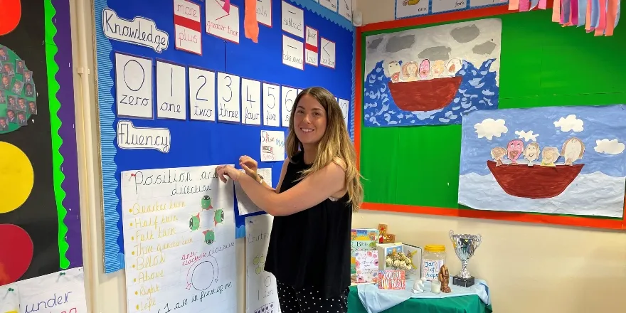 Brunette female in black shirt pins paper to board in classroom.