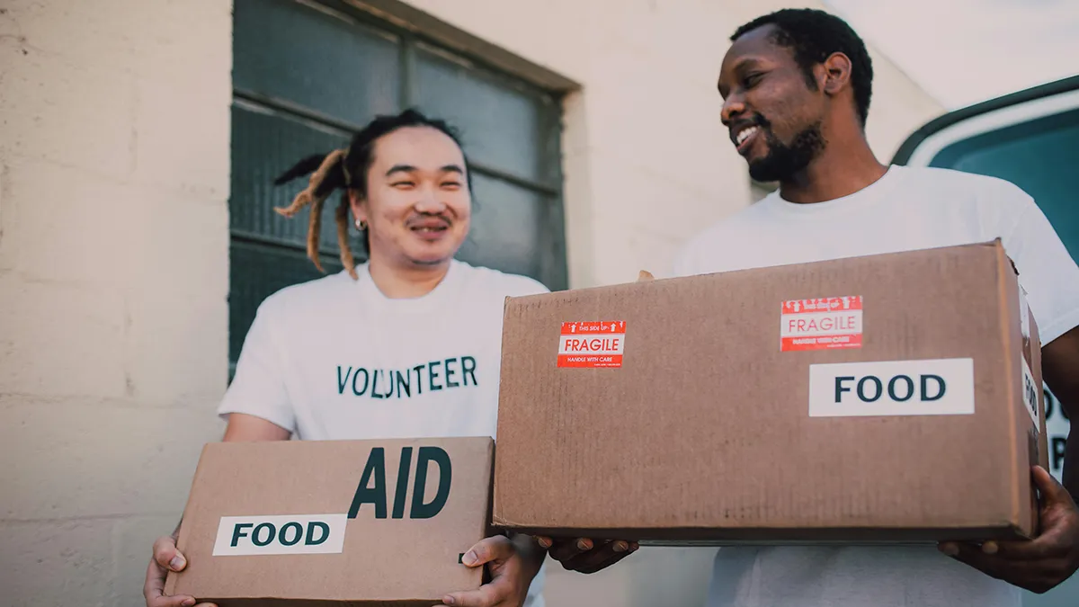 Two men wearing white shirts with the word volunteer on the front and holding boxes of food donations..