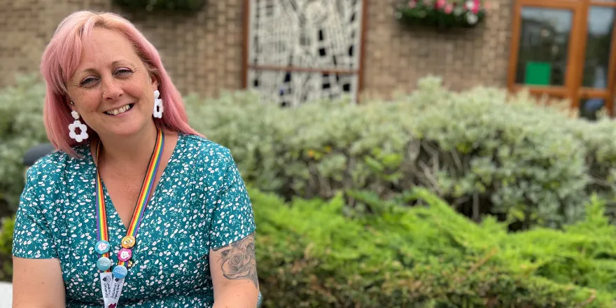 Female with pink hair and green spotty dress sits on bench in front of shrubbery.