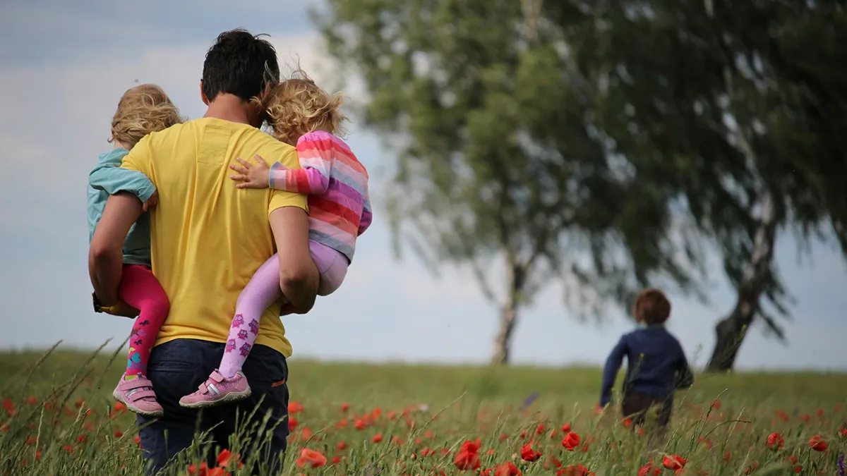 A dad holding two children as a third child runs off ahead, in a field of poppies..