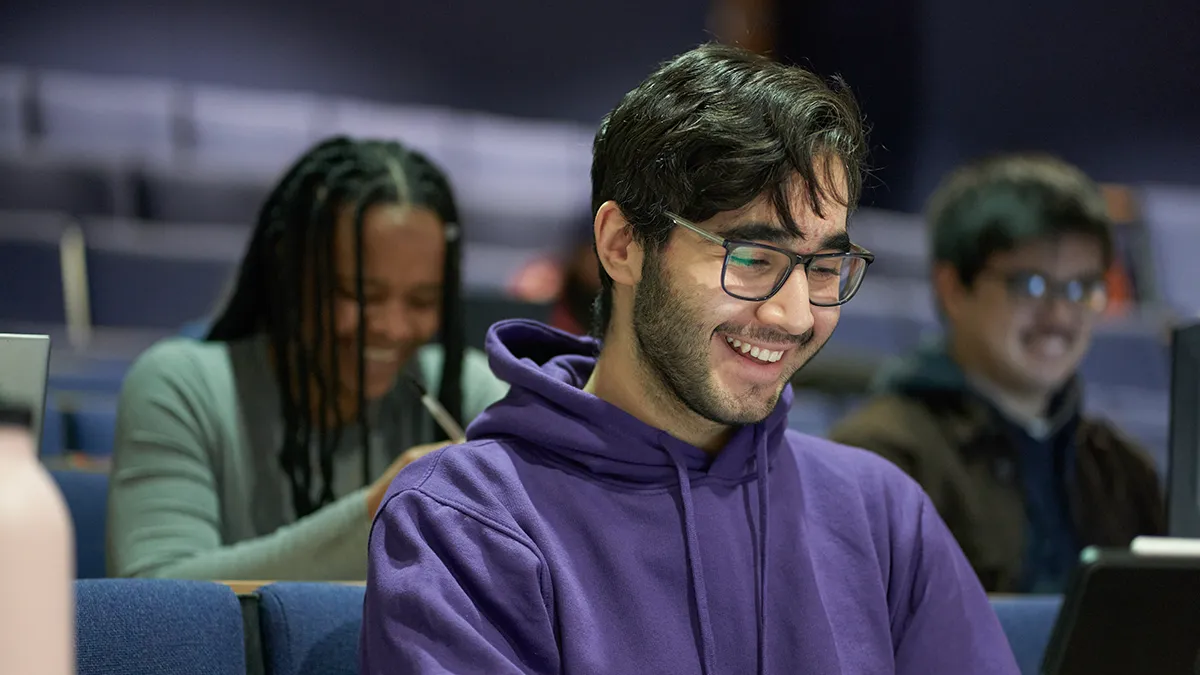 A student in a purple hoodie smiling while looking at a computer in a lecture theatre at Leeds Trinity University..