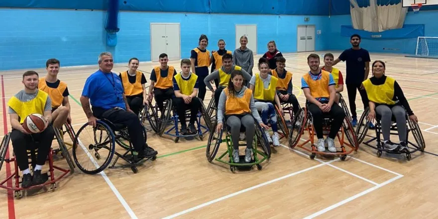 Group of students in wheelchairs and yellow bibs on sports court.