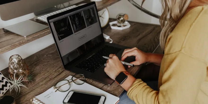 wooden desk with phone, glasses, notebook and hands typing on laptop.