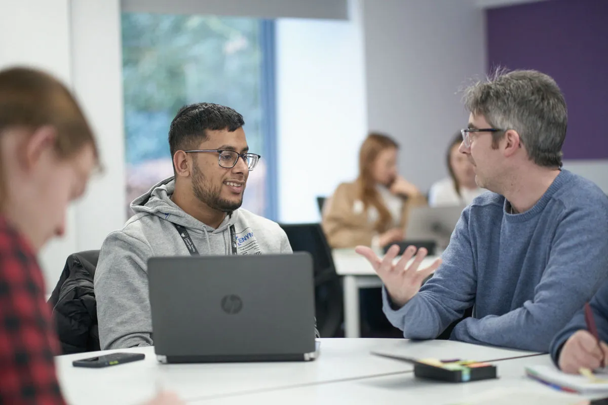 Two men sitting at a table in a classroom, talking and smiling; one has a laptop open while the other gestures with his hand, with other students working in the background..
