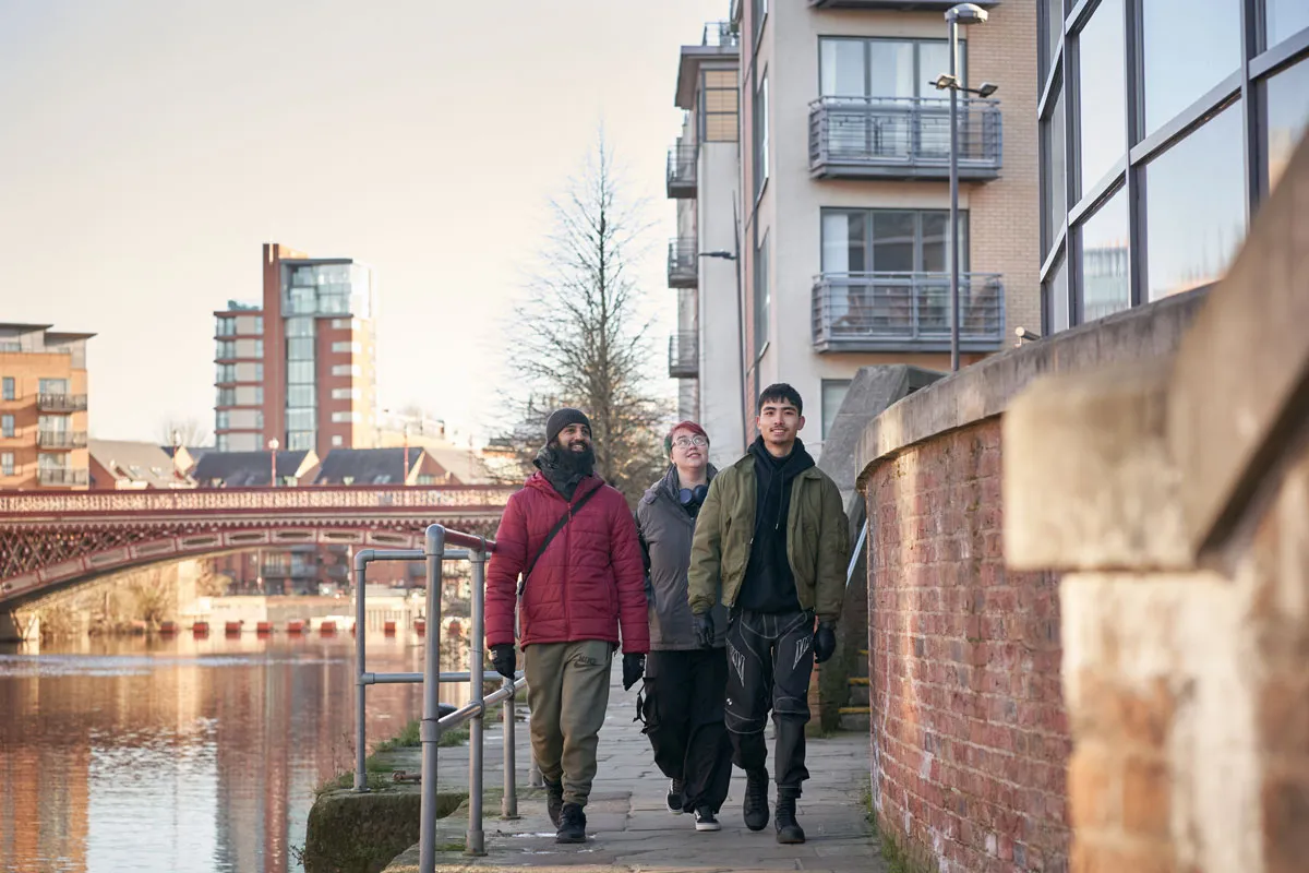 Three students walk along the riverside in Leeds on a sunny day, with modern apartments and a red bridge visible in the background..