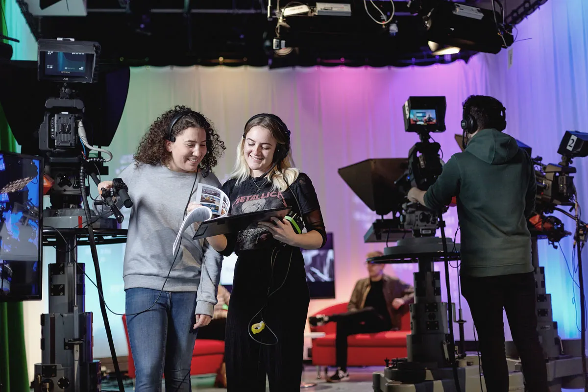 Students working in a TV studio, wearing headsets and operating cameras while reviewing notes, with colourful lighting and a presenter seated on a red sofa in the background..