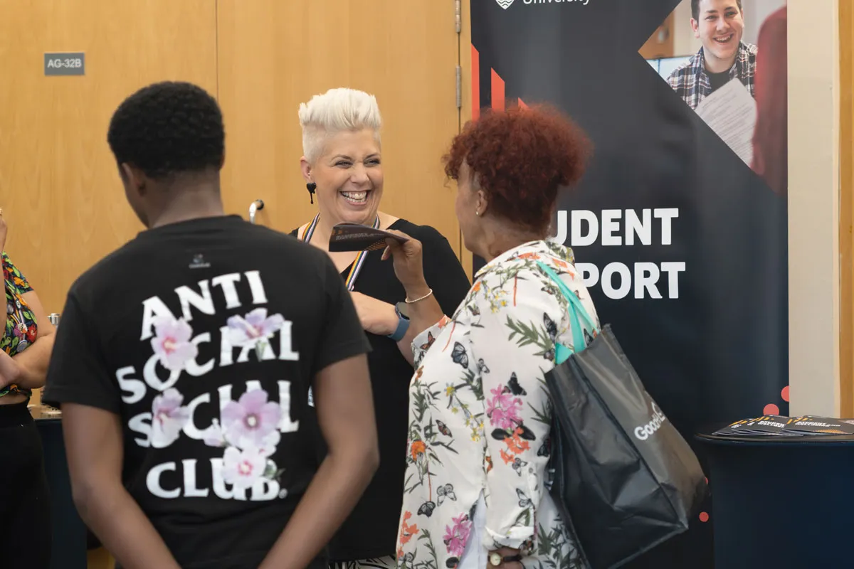 A smiling university staff member talks with visitors at a Student Support stand during an open day, with banners and leaflets visible in the background..