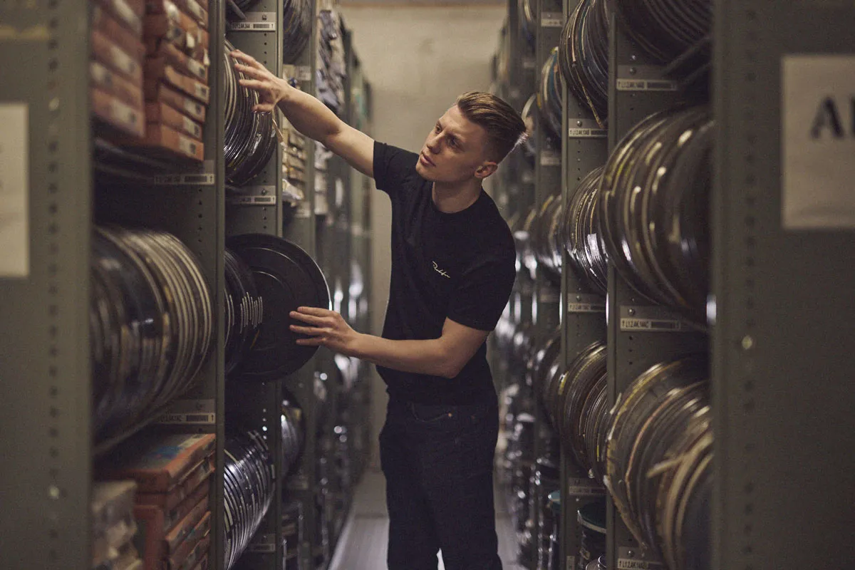 A student stands between shelves filled with film reels, reaching out to select one, in what appears to be an archive or film storage room..