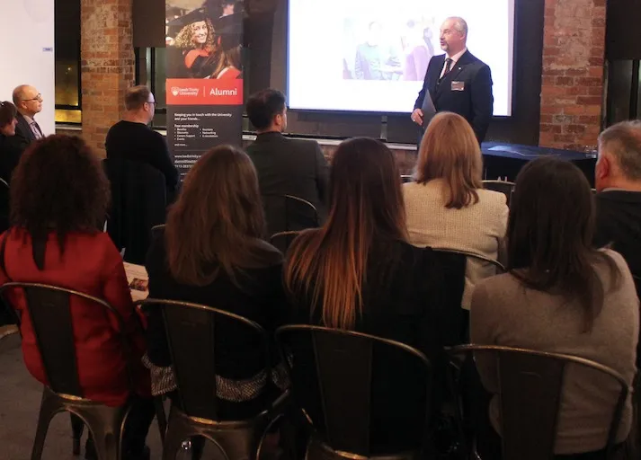 A man in a suit presenting in front of a group of seated guests.