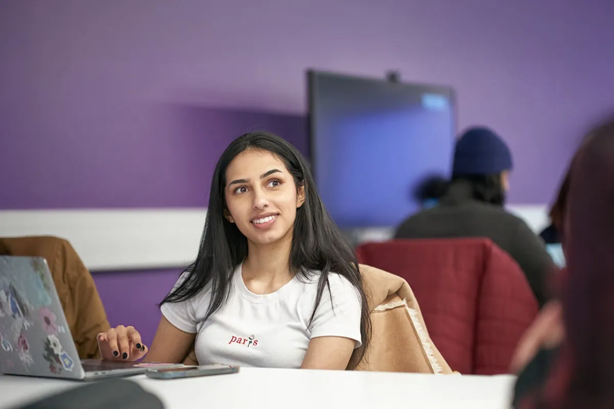 A person sits at a desk using a laptop in a classroom with other people working in the background.