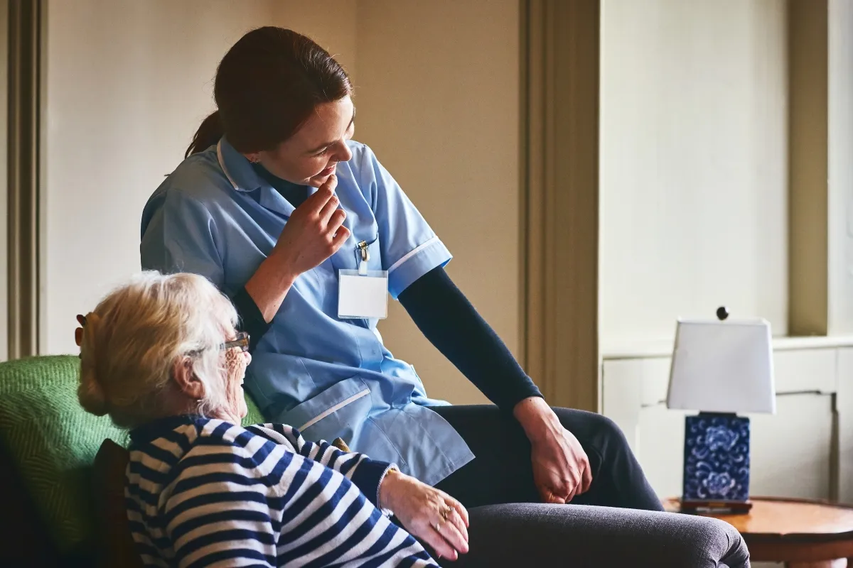 Nurse sitting on the arm of a chair while talking to a patient sat in an arm chair..
