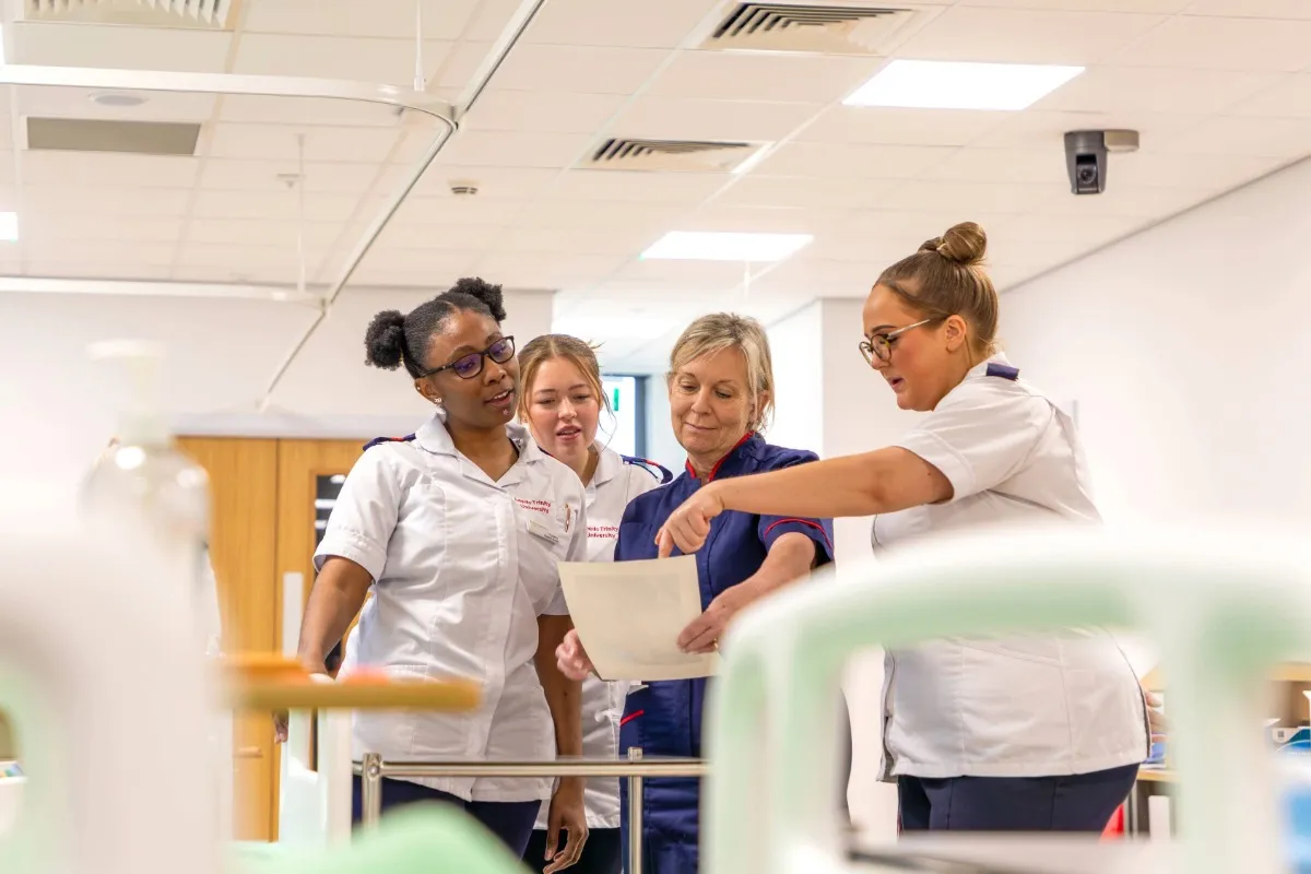 A group of people in clinical uniforms gather around a document in a brightly lit healthcare training room, reviewing information together.