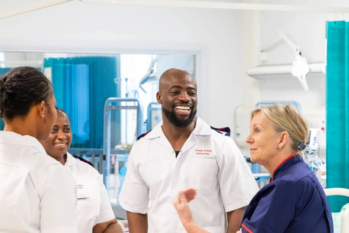 A small group of people wearing clinical uniforms stand together in a healthcare training room while one person speaks and gestures during a discussion.