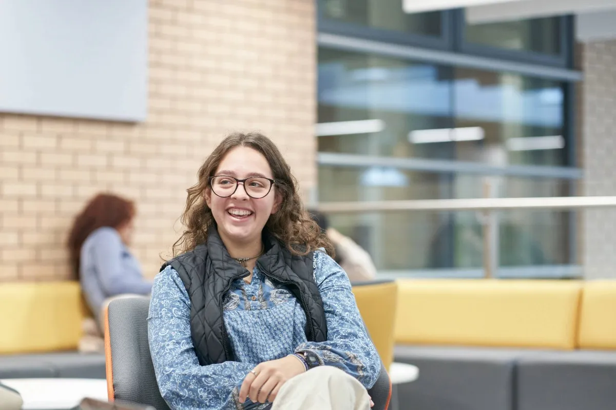 A student sits on a chair holding a notebook, with a bright open space and large windows in the background.