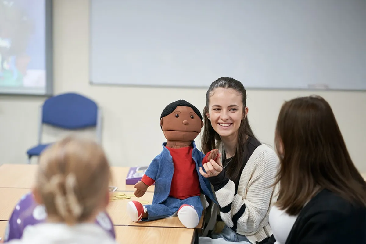 A person sits at a table holding a puppet while speaking to others in a classroom setting.