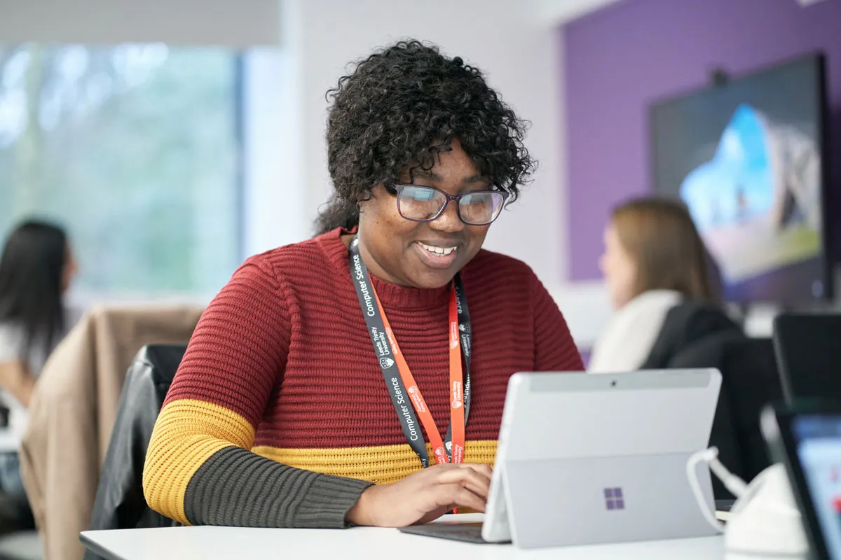 A smiling student works on a Microsoft Surface tablet in a classroom. She is wearing glasses, a red, yellow, and grey striped jumper, and a lanyard labelled 