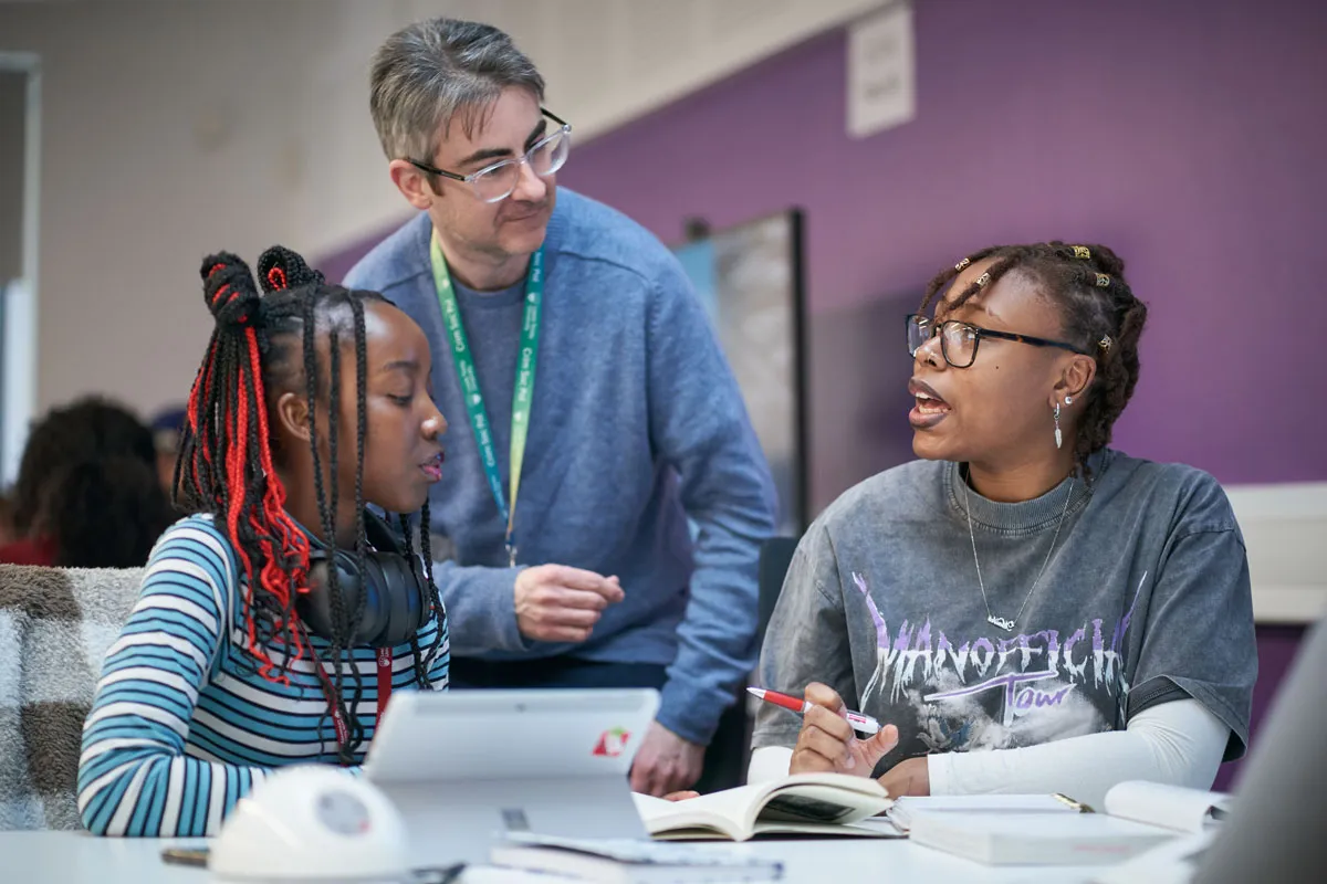 Two students sit at a desk in a classroom, talking and working with open books and a tablet. A lecturer leans over to engage with them. One student wears a striped top and headphones around her neck, the other wears glasses and a dark graphic T-shirt. The background features a purple wall and a screen.