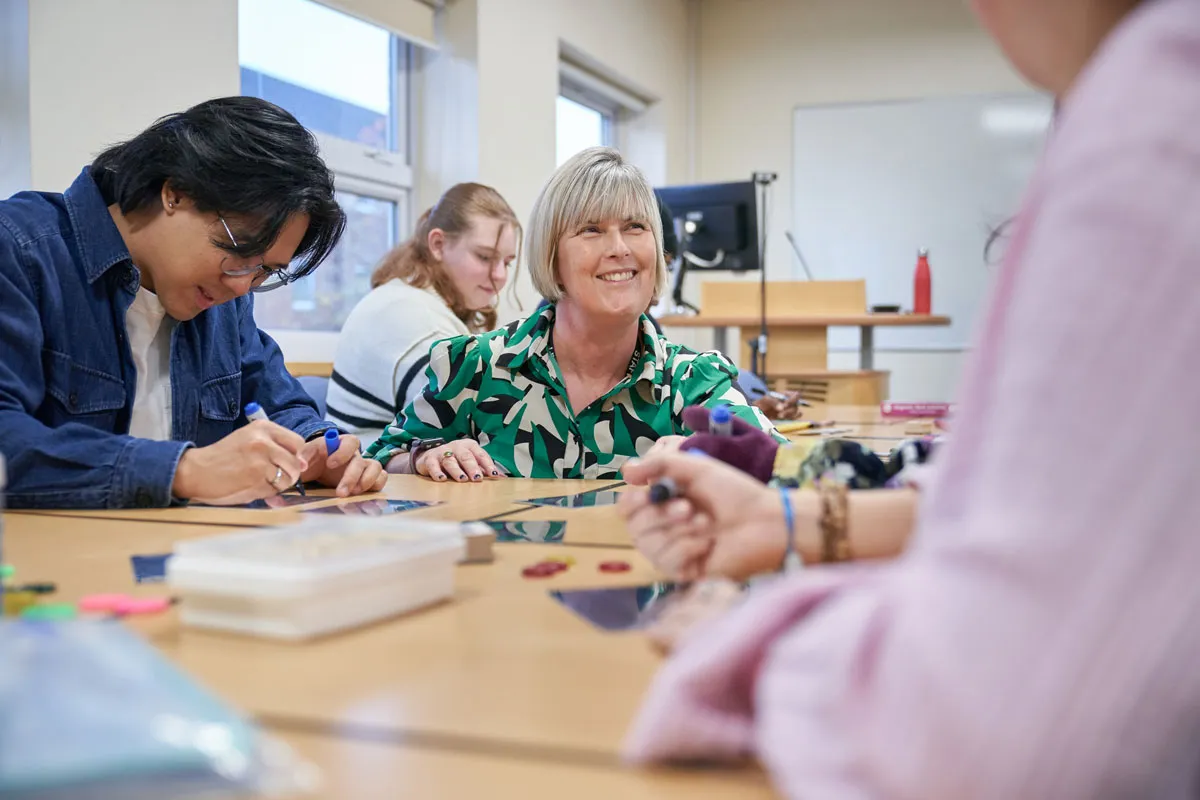 A group of students and a tutor sit around a table in a bright classroom, working on creative tasks. The tutor, wearing a green patterned shirt, smiles while engaging with the group, and materials such as pens and coloured shapes are spread across the table.