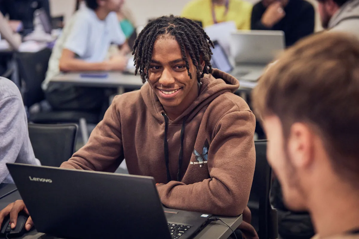 A young man with braids and a brown hoody smiling at someone to his left while using a laptop.