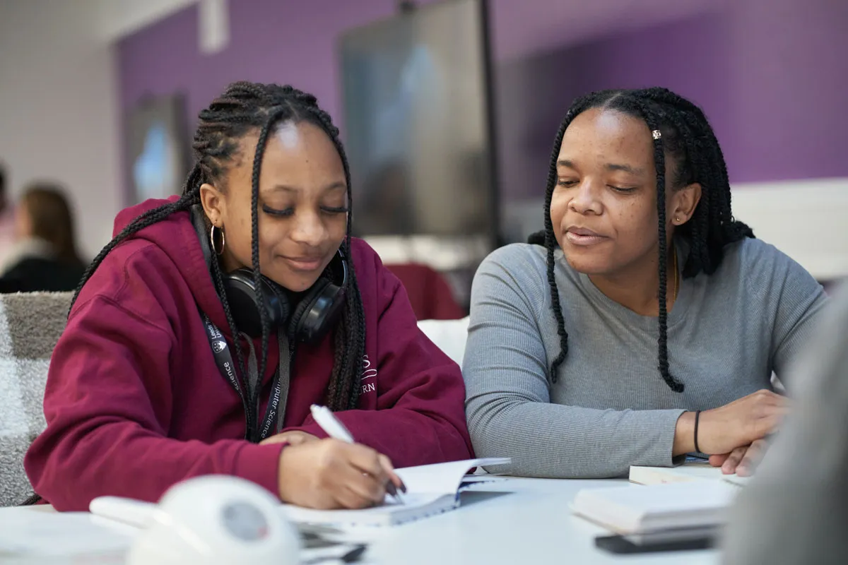 Two students sit at a table working together. One, wearing a maroon hoodie and headphones around her neck, is writing in a notebook while the other, in a grey top, looks on supportively. They appear to be in a study space with a purple wall and screen in the background..