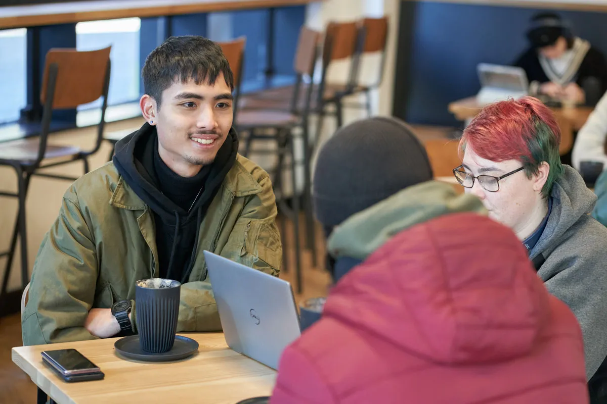 Three students sit around a table in a café, working on laptops and chatting. One student is smiling and engaged in conversation, while another types on their device. Coffee cups and phones are on the table, and other students are visible in the background..