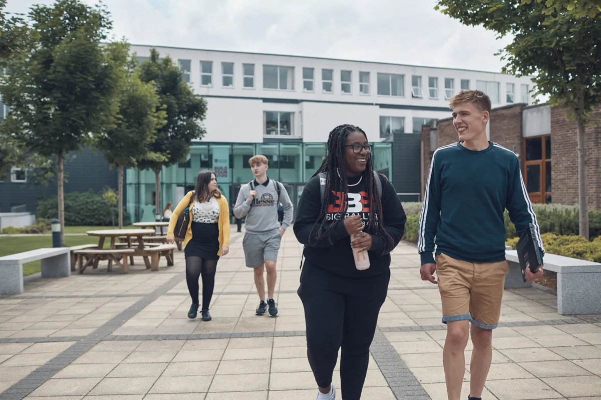 Four university students walk together through a campus courtyard, smiling and chatting. Two students are in the foreground, while two others walk behind them. The area is surrounded by trees, benches, and modern buildings with large windows..