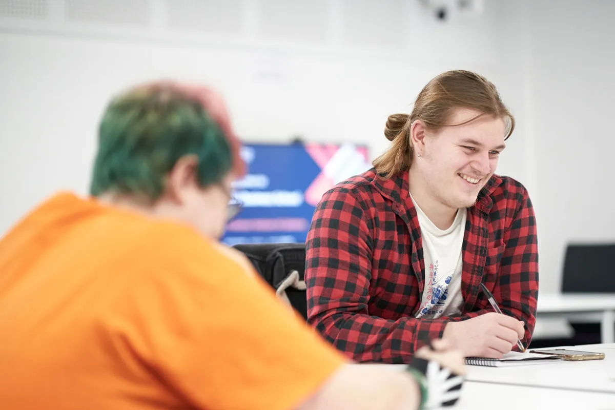 Two students sit at a desk in a brightly lit classroom. One student in a red and black check shirt is smiling while writing in a notebook. The other, with brightly coloured hair and an orange shirt, is slightly out of focus in the foreground..