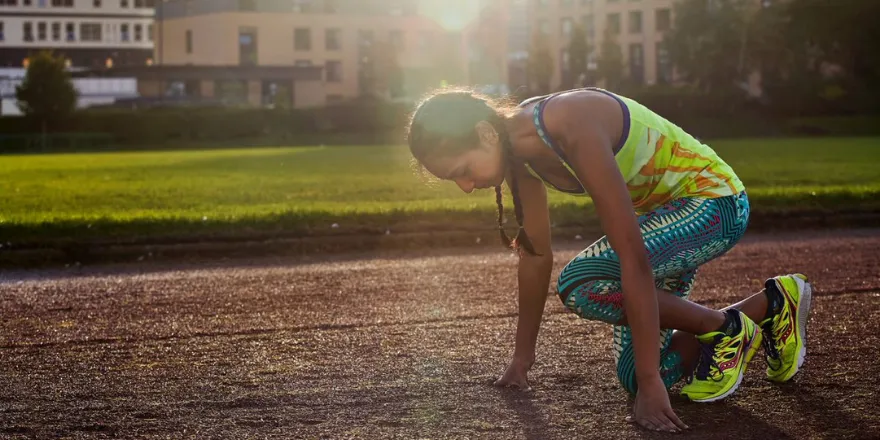 Girl with yellow sports top and blue leggings on crouches on running track in front of University.