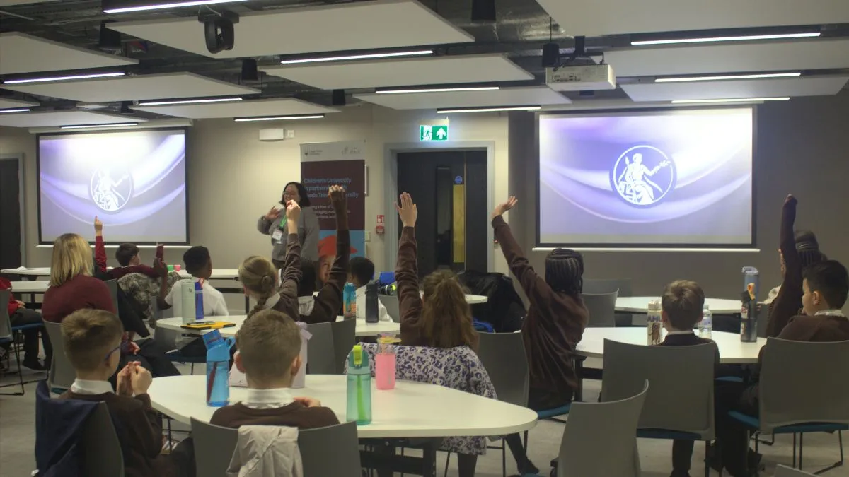A picture of students raising their hands while sat in a classroom.