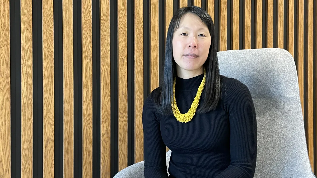A female academic wearing a black top and a yellow necklace, sitting on a grey chair by a wall covered in wooden panels..