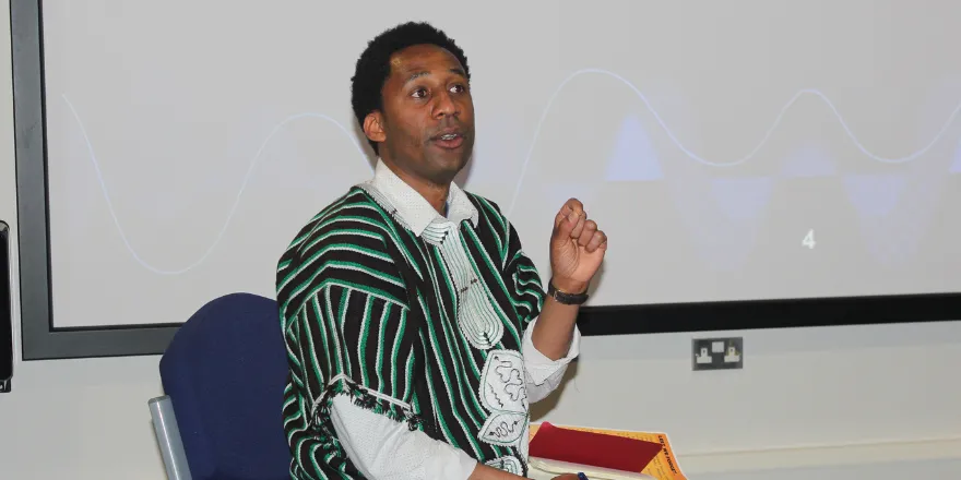 Male sits in front of screen wearing black and green striped shawl.