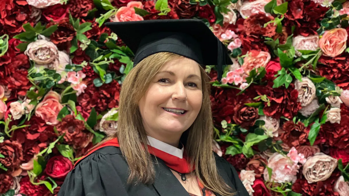 A mature female student posing against a flower wall in graduation cap and gown..