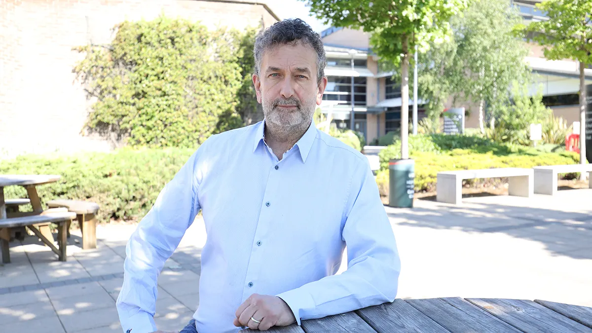 A male academic in a light blue shirt sitting at a wooden table on campus..