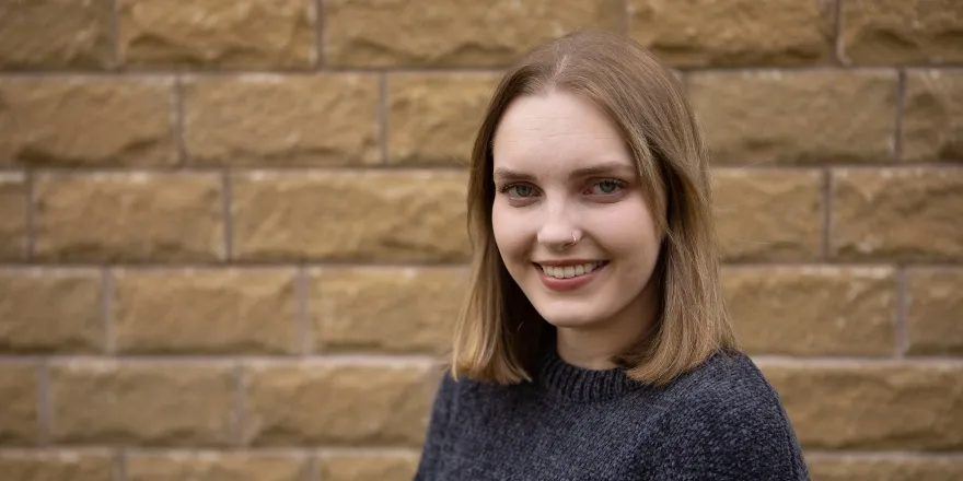 Brunette female in charcoal jumper in front of brick wall.