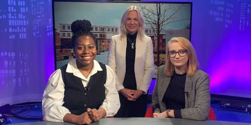 Female students sit at table in front of television screen with female lecturer stood up in middle.
