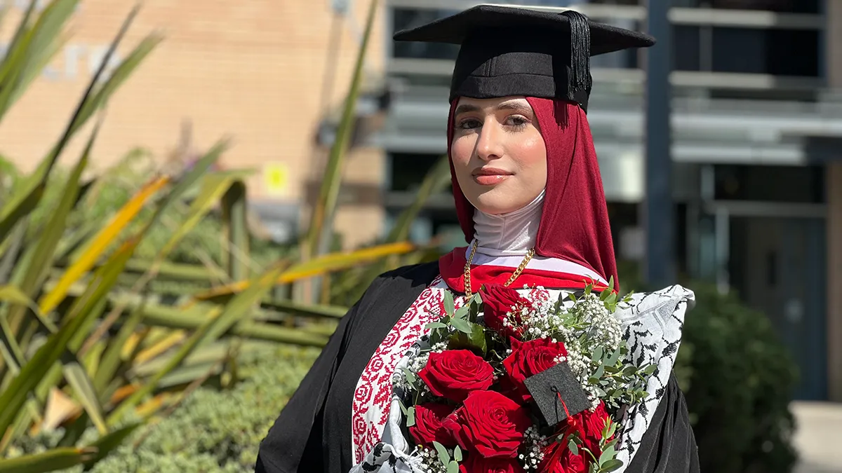 A student in a burgundy hijab, and Graduation cap and gown, holding a bouquet of red roses and smiling..
