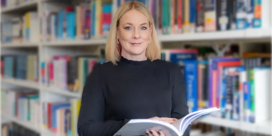 Blonde female stands holding open book in front of bookcase.