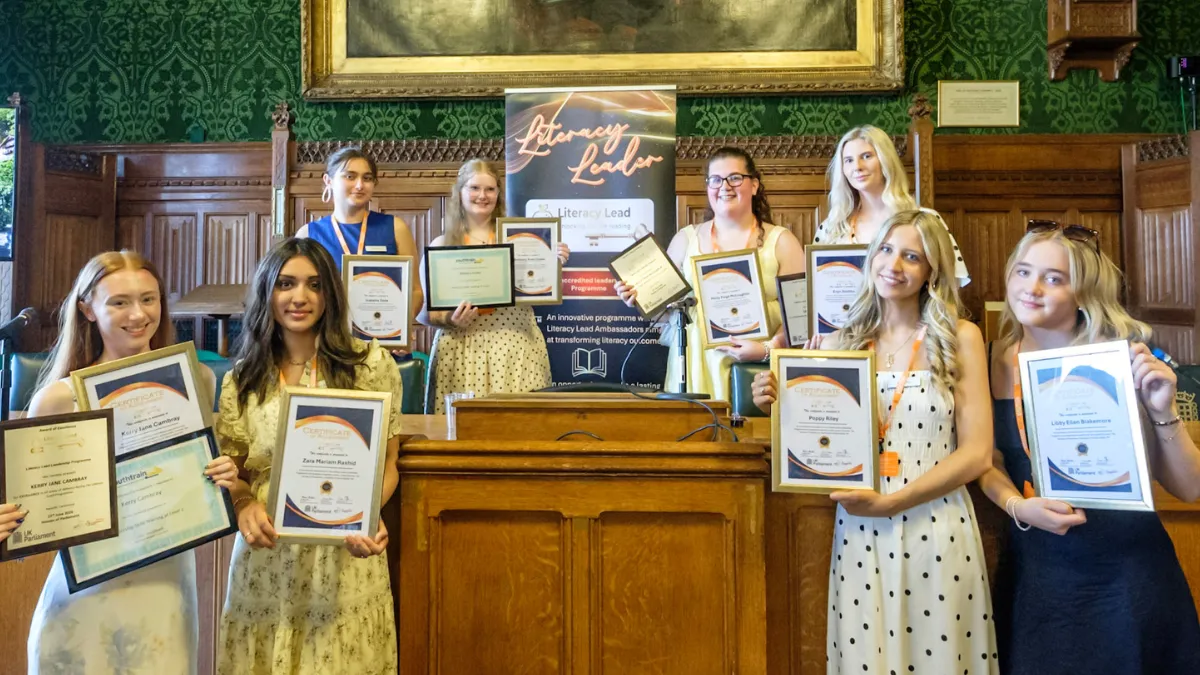 A group of teacher education students from Leeds Trinity University posing in Parliament with certificates accrediting the Literacy Leads project they took part in. A banner with the title Literacy Leads is behind them and they are gathered around a wooden desk..