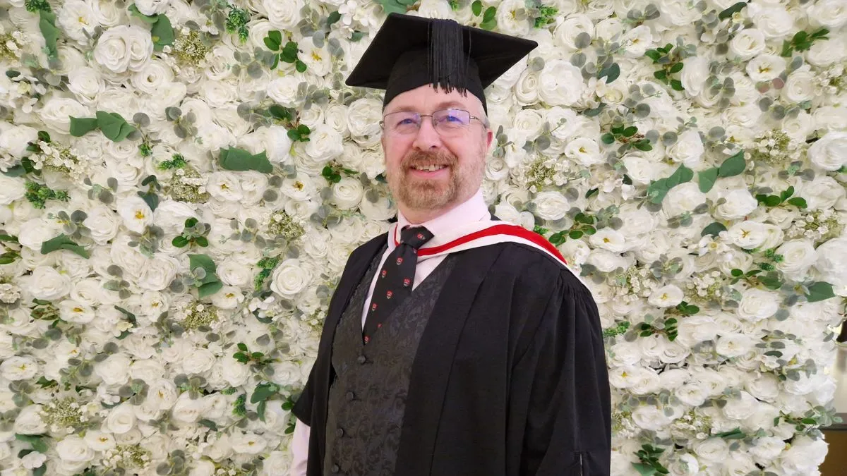 A graduate wearing a cap and gown stands in front of a white flower wall.