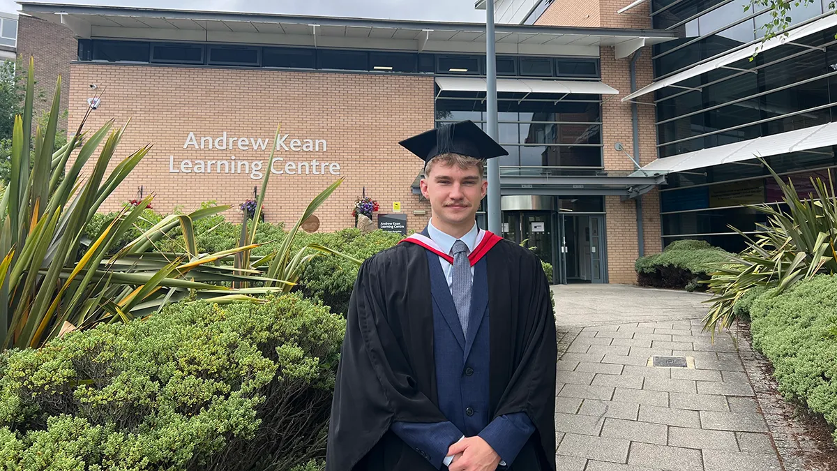 A student in graduation cap and gown smiling in front of the Andrew Kean Learning Centre at Leeds Trinity University..
