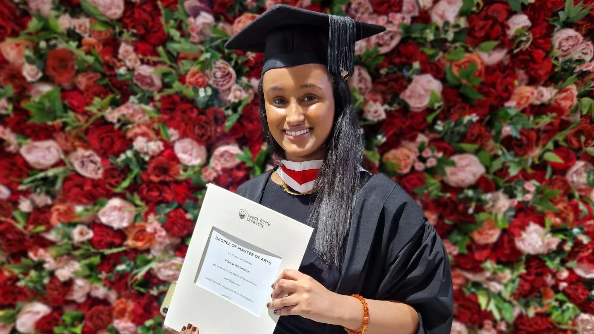 A girl in a graduation gown holds her certificate in front of a red and pink wall of flowers.